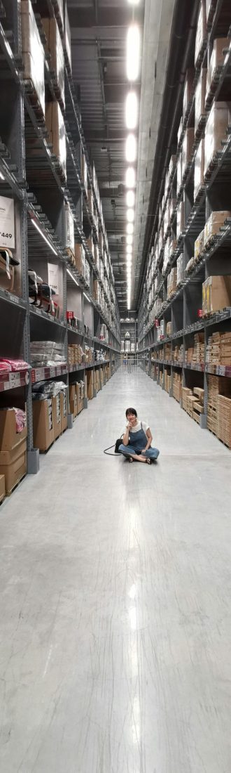 An Asian woman sits in a large warehouse aisle filled with inventory racks and shelves.